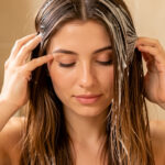 Jeune femme appliquant délicatement un masque nourrissant sur ses cheveux, avec des racines brillantes et des longueurs sèches abîmées par la chaleur.